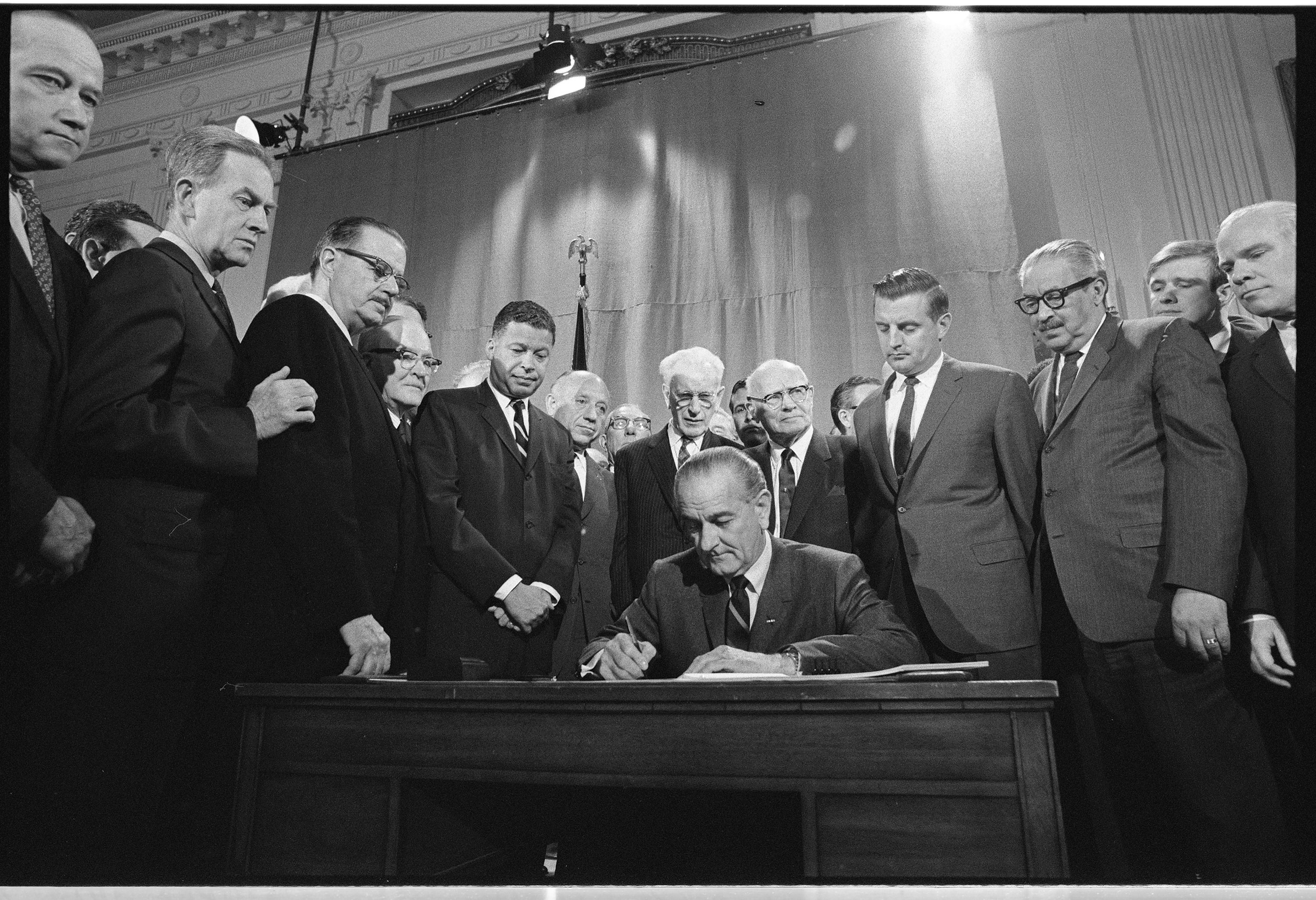 President Johnson, seated at desk, signs a piece of paper while a crowd of men in suits observe. 